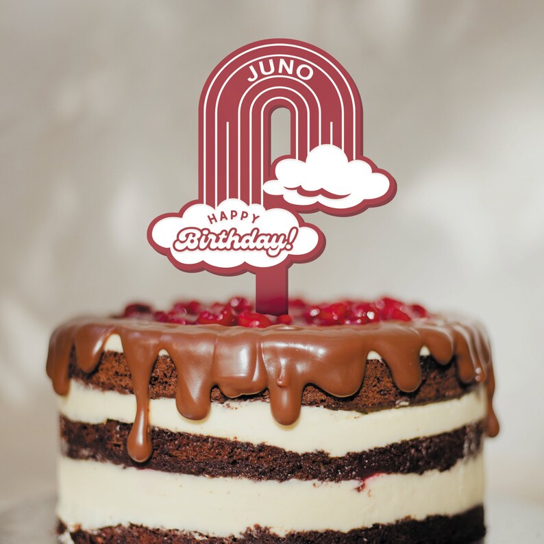 Op de afbeelding: Een chocoladetaart met witte frosting en rode sprinkles, bedekt met een rode en witte regenboogtaarttopper met de tekst "JUNO" en "Happy Birthday!"