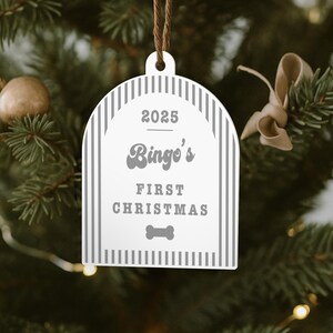 May include: White Christmas ornament with gray vertical stripes and the text "2025 Bingo's First Christmas" and a dog bone graphic. The ornament hangs from a brown string against a blurred Christmas tree background.