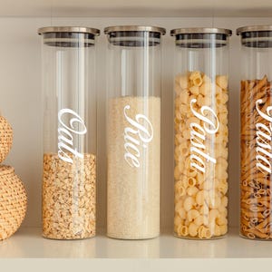 May include: Four clear glass canisters with silver lids, filled with oats, rice, pasta, and pasta. Each canister has a white label with the name of the food written in cursive.