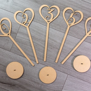 May include: Five wooden heart-shaped table number holders with numbers 1, 2, 3, 4, and 5. Each holder has a wooden stick and a round base.