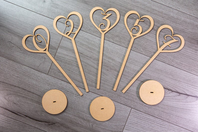 May include: Five wooden heart-shaped table number holders with numbers 1, 2, 3, 4, and 5. Each holder has a wooden stick and a round base.