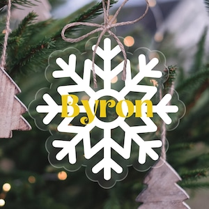 Op de afbeelding: Witte sneeuwvlok ornament met de naam "Byron" in gele letters. De ornament hangt aan een kerstboom.