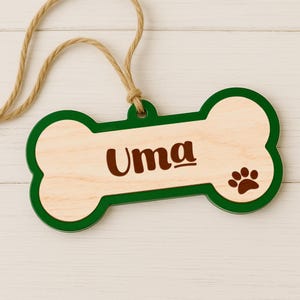 May include: A wooden dog bone-shaped tag with the name "Uma" in brown lettering. The tag has a green border and a paw print on the right side. A natural twine cord is attached for hanging.