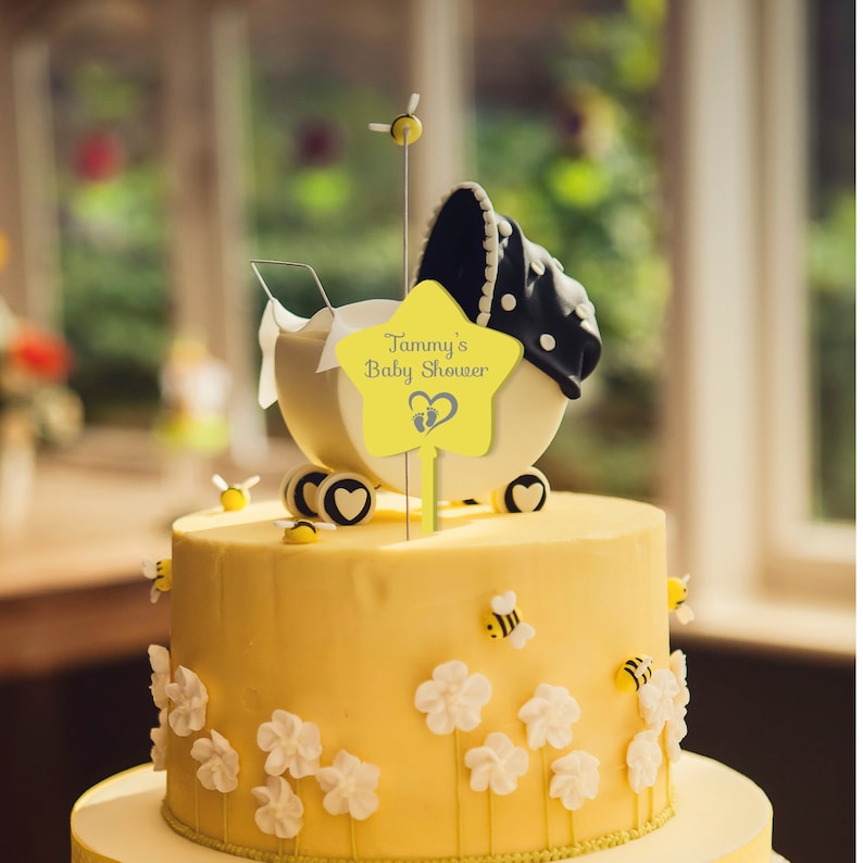 May include: Yellow and white two-tiered cake decorated with white flowers, bees, and a yellow star topper that says "Tammy's Baby Shower" with a baby footprint and heart graphic.