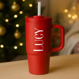 May include: A red insulated tumbler with a handle and a white straw. The word "LUCY" is printed vertically in white on the side of the tumbler. The tumbler is sitting on a wooden surface.