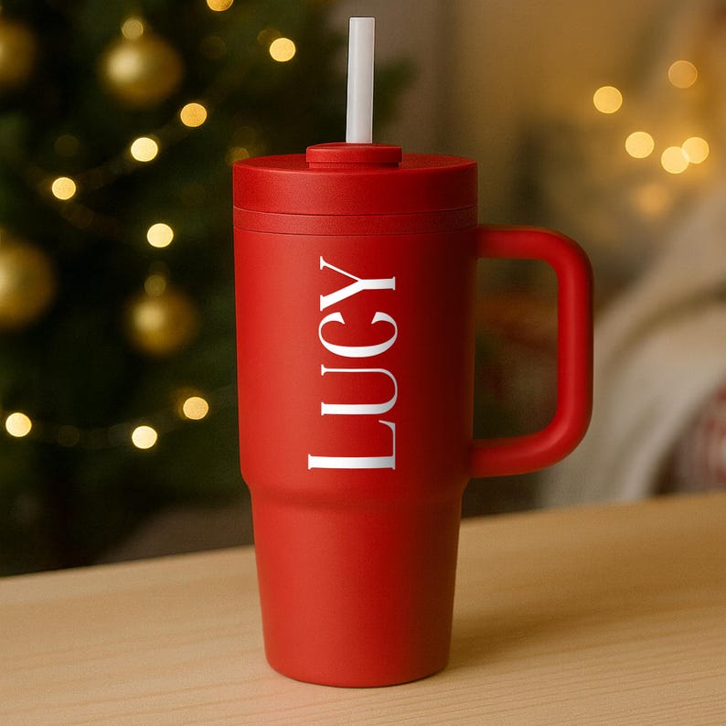May include: A red insulated tumbler with a handle and a white straw. The word "LUCY" is printed vertically in white on the side of the tumbler. The tumbler is sitting on a wooden surface.