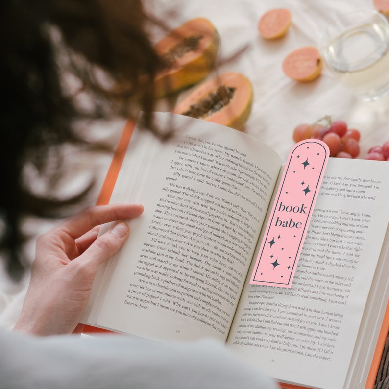 May include: A pink bookmark with the text "book babe" and pink stars. The bookmark is placed in a book with a white background.