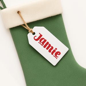 May include: A green Christmas stocking with a white cuff and a white tag. The tag is attached with twine and has the name "Jamie" printed in red. The stocking is made of a textured fabric.
