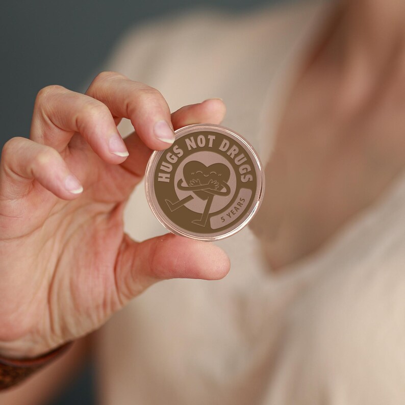 May include: A round, clear plastic token with a brown design. The design features a heart with two figures hugging inside. The text "Hugs Not Drugs" is written around the heart. The text "5 Years" is written below the heart.
