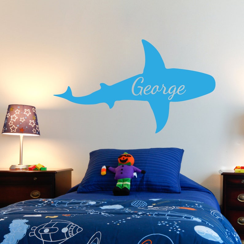 May include: A blue shark silhouette wall decal with the name "George" written on it. The decal is on a white wall above a blue and white patterned bed with a stuffed toy on it.