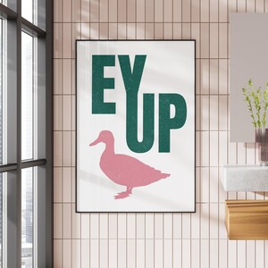 May include: A white poster with the text "EY UP" in dark green letters. A pink silhouette of a duck is below the text.