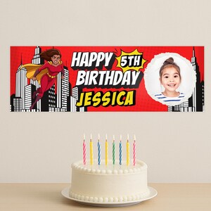 Puede incluir: Una pancarta roja con el texto "HAPPY 5TH BIRTHAY JESSICA" y una superhero&iacute;na de dibujos animados. Una foto de una ni&ntilde;a peque&ntilde;a est&aacute; en un c&iacute;rculo blanco. Una tarta blanca con velas est&aacute; delante de la pancarta.