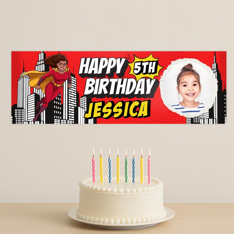 Puede incluir: Una pancarta roja con el texto "HAPPY 5TH BIRTHAY JESSICA" y una superhero&iacute;na de dibujos animados. Una foto de una ni&ntilde;a peque&ntilde;a est&aacute; en un c&iacute;rculo blanco. Una tarta blanca con velas est&aacute; delante de la pancarta.