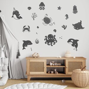 Op de afbeelding: Een set zwarte en witte muurstickers met zeedieren. De stickers bevatten een narwal, een schildpad, een octopus, een krab, een dolfijn, een zeepaardje, een kwal, een ster en een rog. De stickers zijn perfect om een kinderkamer een vleugje whimsy te geven.