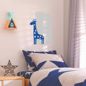 May include: A blue giraffe wall decal with white spots, standing next to a light blue letter R on a white wall. The decal is above a bed with a blue and white star patterned duvet cover.