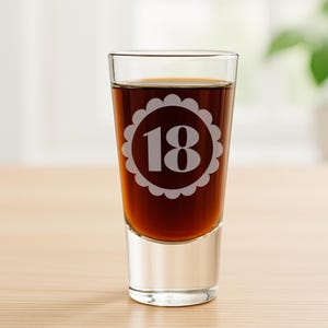 May include: A clear shot glass filled with a dark liquid. The glass has a white scalloped design with the number 18 in the center. The glass is sitting on a light brown surface.