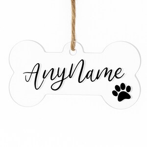 以下が含まれることがあります： 白い骨型のペットタグ。黒い筆記体で「AnyName」と黒い足跡が描かれています。タグは天然の麻ひもで吊り下げられています。