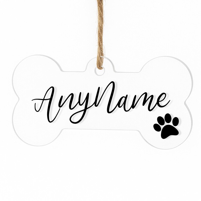 以下が含まれることがあります： 白い骨型のペットタグ。黒い筆記体で「AnyName」と黒い足跡が描かれています。タグは天然の麻ひもで吊り下げられています。