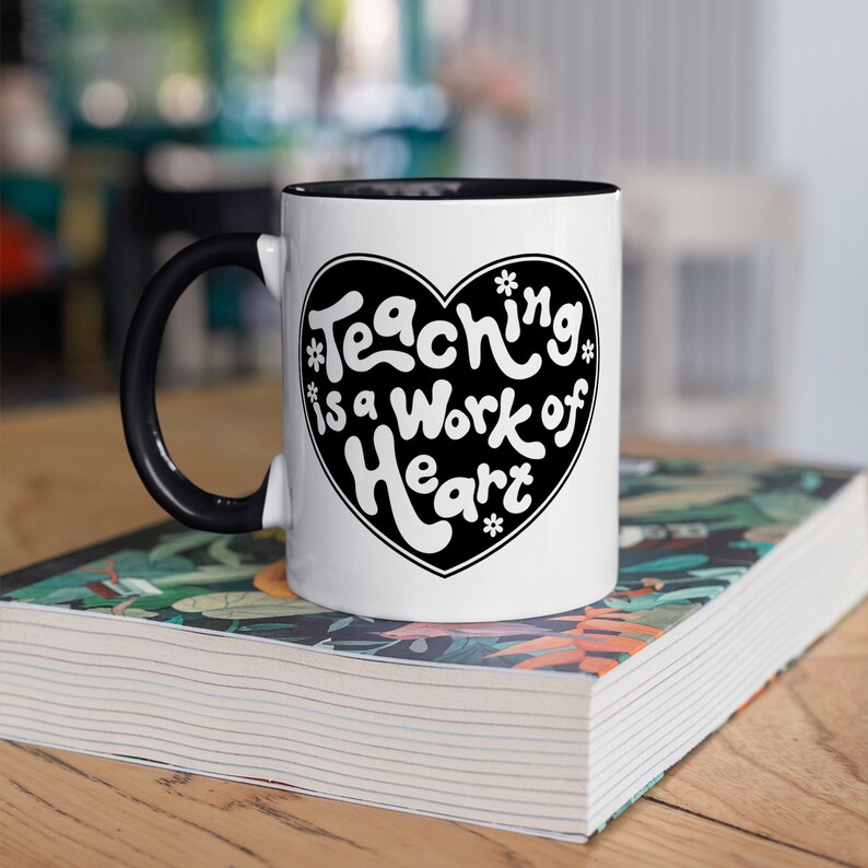 Puede incluir: Taza de cer&aacute;mica blanca con asa y borde negros. La taza tiene un dise&ntilde;o de coraz&oacute;n negro con el texto "Teaching is a work of heart" en blanco.