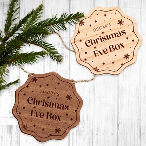 May include: Two wooden Christmas Eve box decorations with a rustic wood grain finish. The top decoration is light brown with the text "OSCAR'S Christmas Eve Box" in brown lettering. The bottom decoration is dark brown with the text "MADDY'S Christmas Eve Box" in brown lettering. Both decorations have a snowflake design.