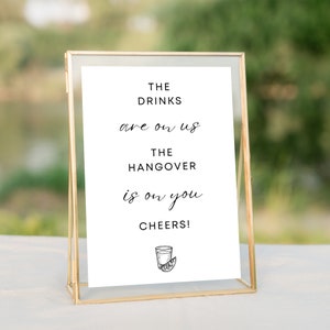 May include: A white sign with black text that reads "The drinks are on us. The hangover is on you. Cheers!" with a drawing of a margarita glass with a lime wedge. The sign is in a gold frame.