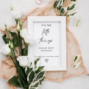 May include: White framed sign with the text "It's the little things. Please help yourself to whatever you may need." with a small heart symbol at the bottom. The sign is surrounded by white flowers and green leaves.