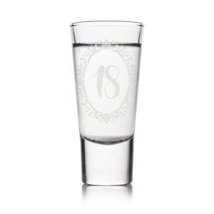May include: A clear shot glass with the number 18 etched in a decorative oval frame. The glass is filled with a clear liquid and is suitable for a birthday celebration. The glass is on a white background.