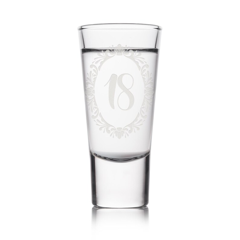 May include: A clear shot glass with the number 18 etched in a decorative oval frame. The glass is filled with a clear liquid and is suitable for a birthday celebration. The glass is on a white background.