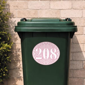 May include: A dark green trash bin with a pink and white oval sticker. The sticker features the number 208 in white, surrounded by a decorative floral pattern. The bin is set against a brick wall.