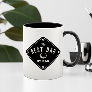 May include: White ceramic coffee mug with a black rim and handle. The mug features a black and white graphic of a golf ball and the text "The Best Dad By Par".