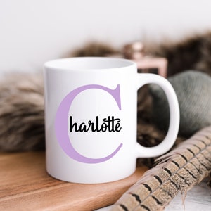 K&ouml;nnte beinhalten: Wei&szlig;e Keramiktasse mit einem violetten Buchstaben C und dem Wort "charlotte" in Schwarz innerhalb des Buchstabens.