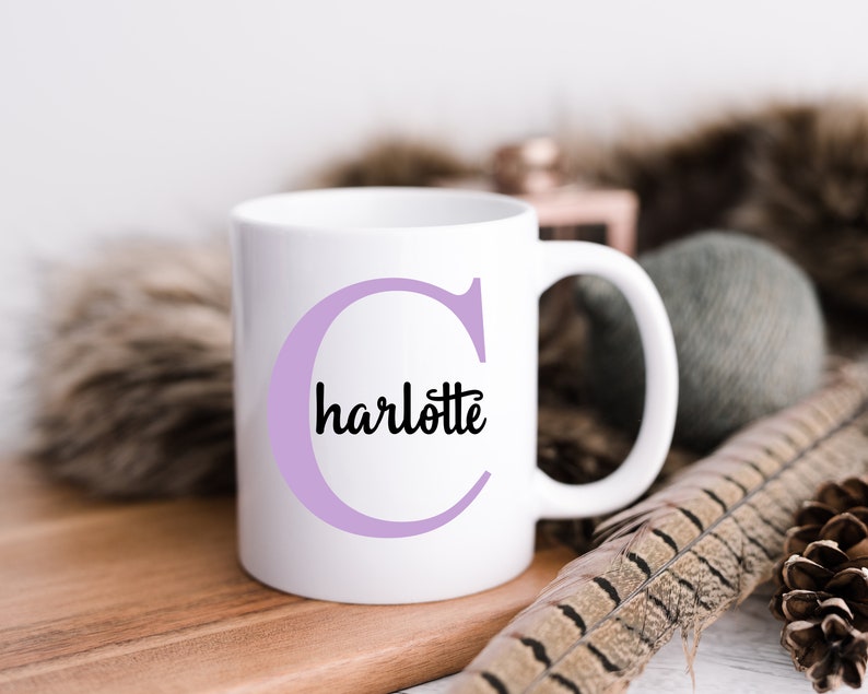 K&ouml;nnte beinhalten: Wei&szlig;e Keramiktasse mit einem violetten Buchstaben C und dem Wort "charlotte" in Schwarz innerhalb des Buchstabens.