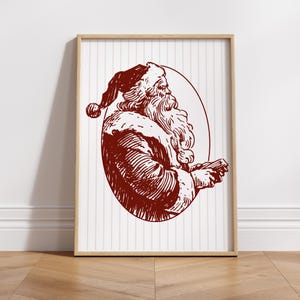 May include: A framed print featuring a vintage illustration of Santa Claus in a burgundy color. The artwork depicts Santa in profile, holding a small object, set against a white background with vertical stripes. The frame is a light wood color.