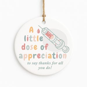May include: A round, white ceramic ornament with a syringe illustration and the text "A little dose of appreciation to say thanks for all you do!" The syringe is red and light blue.