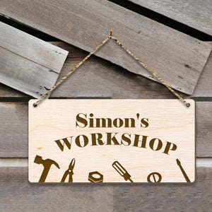 May include: A wooden sign with the text "Simon's Workshop" in brown lettering. The sign has a rope for hanging and features a hammer, pliers, a nut, a screwdriver, and a wrench.