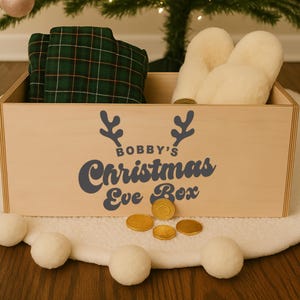 May include: A wooden Christmas Eve box with the text "Bobby's Christmas Eve Box" in dark blue. The box contains green plaid pajamas and cream-colored slippers. Gold chocolate coins and white pom-poms are in front of the box.