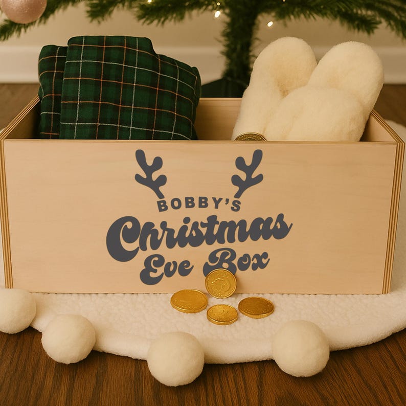 May include: A wooden Christmas Eve box with the text "Bobby's Christmas Eve Box" in dark blue. The box contains green plaid pajamas and cream-colored slippers. Gold chocolate coins and white pom-poms are in front of the box.
