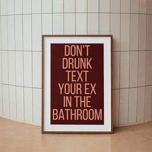 Op de afbeelding: Een ingelijst print met een bordeauxrode achtergrond en een witte rand. De tekst "DON'T DRUNK TEXT YOUR EX IN THE BATHROOM" is in oranje geschreven.