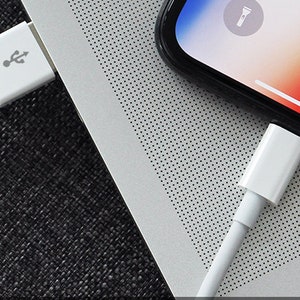 Op de afbeelding: Twee witte Lightning-naar-USB-kabels aangesloten op een zilveren laptop en een zwarte smartphone. Het smartphone-scherm toont een zaklamppictogram.