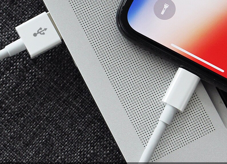 Op de afbeelding: Twee witte Lightning-naar-USB-kabels aangesloten op een zilveren laptop en een zwarte smartphone. Het smartphone-scherm toont een zaklamppictogram.