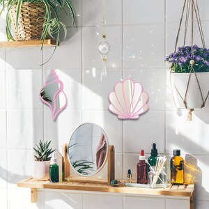 May include: Two pink seashell-shaped mirrors hang on a white tiled wall above a wooden shelf with a round mirror, a potted succulent, and various bottles and jars.