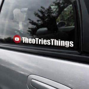 May include: A white decal on a car window with the text "TheoTriesThings" in a bold, sans-serif font. The decal includes a red circle with a white play button, resembling a YouTube logo. The car is silver.