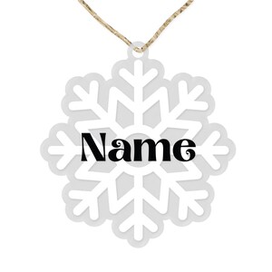 Op de afbeelding: Witte sneeuwvlok ornament met een touwtje om op te hangen. De sneeuwvlok heeft een witte omtrek en een grijs centrum. Het woord "Naam" is in het zwart in het midden van de sneeuwvlok gedrukt.
