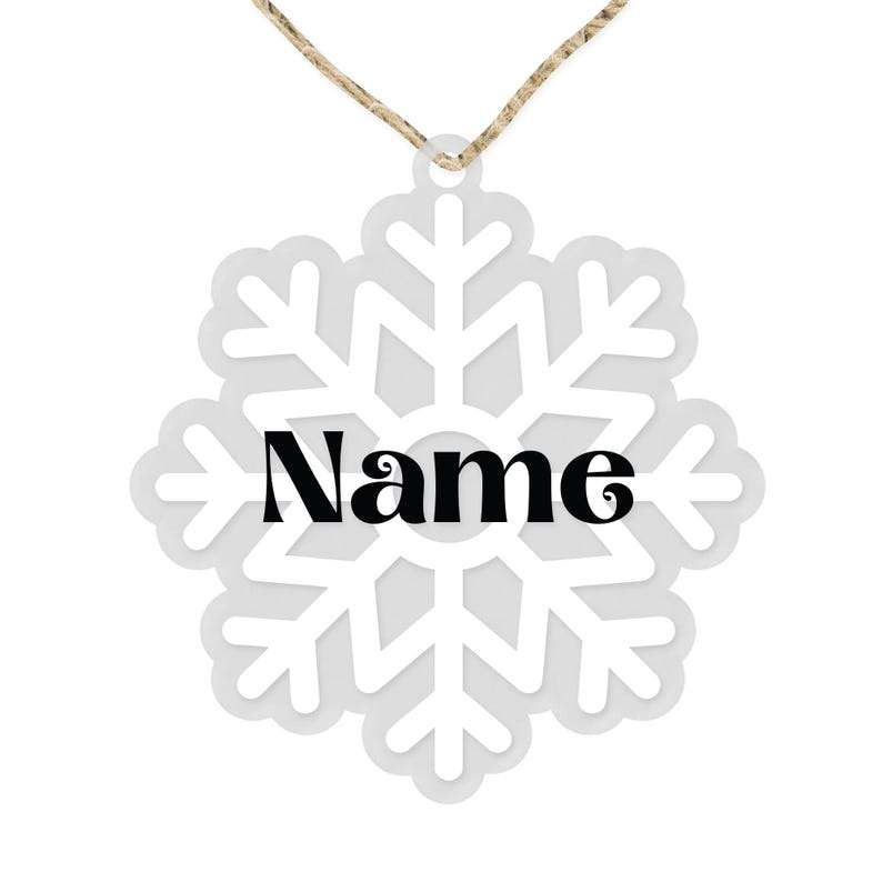 Op de afbeelding: Witte sneeuwvlok ornament met een touwtje om op te hangen. De sneeuwvlok heeft een witte omtrek en een grijs centrum. Het woord "Naam" is in het zwart in het midden van de sneeuwvlok gedrukt.