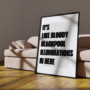 May include: A framed print with the text "IT'S LIKE BLOODY BLACKPOOL ILLUMINATIONS IN HERE" in bold black letters. The print is in a black frame and leans against a beige sofa. The room has a wooden floor.