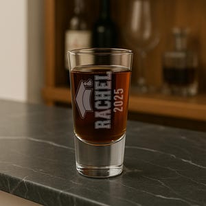 May include: A clear shot glass filled with a dark amber liquid. The glass is etched with the name "RACHEL", a graduation cap, and the year "2025". The glass is sitting on a dark gray surface.