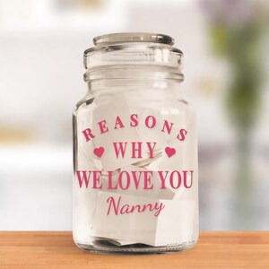 May include: A clear glass jar with a lid, filled with white paper notes. The jar has pink text that reads "REASONS WHY WE LOVE YOU Nanny" with heart symbols. The jar sits on a wooden surface.
