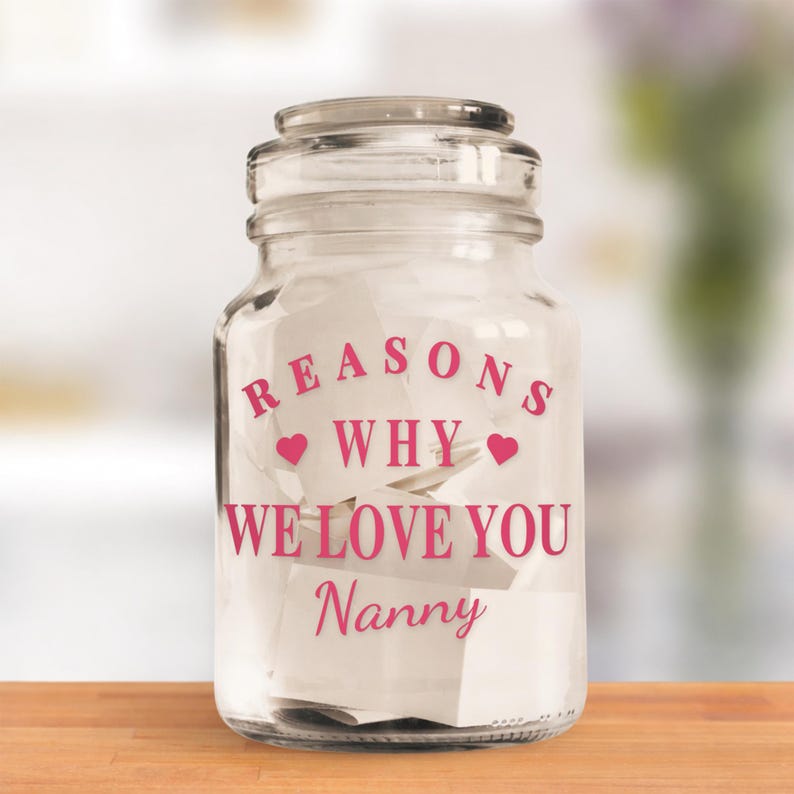 May include: A clear glass jar with a lid, filled with white paper notes. The jar has pink text that reads "REASONS WHY WE LOVE YOU Nanny" with heart symbols. The jar sits on a wooden surface.