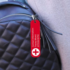 Op de afbeelding: Een rode rechthoekige sleutelhanger met witte tekst en een wit kruissymbool. De sleutelhanger is bevestigd aan een zwarte gewatteerde rugzak. De tekst op de sleutelhanger luidt "Contact Information: Tracey (Mum) 07352618792".