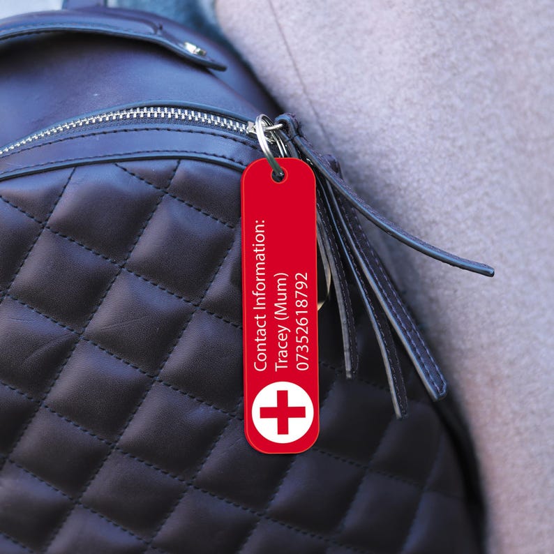 Op de afbeelding: Een rode rechthoekige sleutelhanger met witte tekst en een wit kruissymbool. De sleutelhanger is bevestigd aan een zwarte gewatteerde rugzak. De tekst op de sleutelhanger luidt "Contact Information: Tracey (Mum) 07352618792".